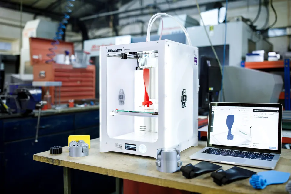 Stolní 3D tiskárna tiskne prototyp v dílně vedle notebooku se slicovacím softwarem a hotových vytištěných dílů.