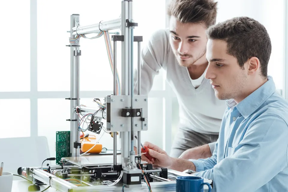 Dva lidé pracují u stolní 3D tiskárny v domácí dílně.