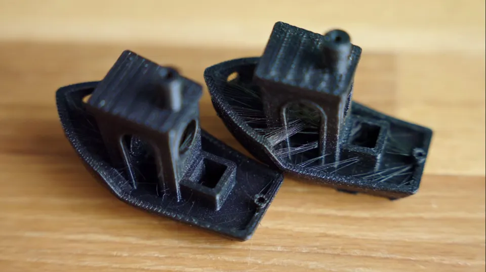 Dva černé 3D tisky Benchy s viditelným stringingem a nerovným povrchem způsobeným problematickým filamentem